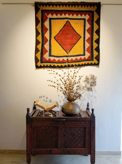 A decorative rug on wall hangs above an antique wooden chest with an open book and a vase of dried flowers - Beautiful Homes